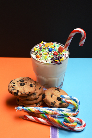 Glass of milk shake with cream, sprinkles, colorful dragee, marmalade jelly candies inside, chocolate chip cookies, striped caramel candies and purple ribbon on colorful black orange blue backgroundの写真素材