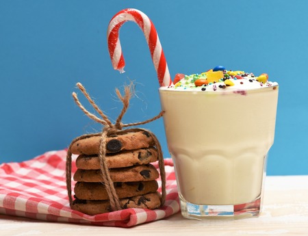 Glass of milk shake with cream, sprinkles, striped caramel candy, colorful dragee, marmalade jelly candies inside, chocolate chip cookies with string and checkered pink napkin on blue white backgroundの写真素材