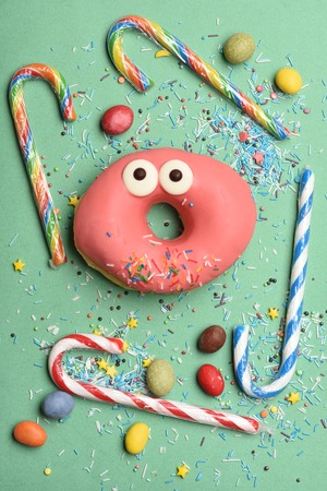 Pink funny surprised glazed donut with sprinkles, striped caramel candies, colorful dragee with...