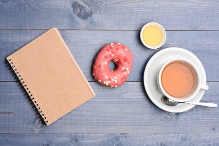 Pink glazed donut with icing or sprinkles, honey, cup with tea with spoon on plate and notepad on blue vintage wooden backgroundの写真素材
