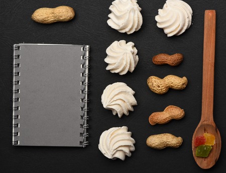 White marshmallow, recipe book or notepad, peanuts, marmalade or jelly candies and wooden spoon on black textured background, homemade cooking concept, copy spaceの写真素材