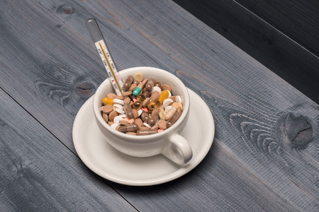 Medical capsules, tablets and clinical thermometer inside coffee cup on vintage wooden background....
