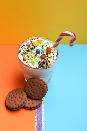 Glass of milk shake with cream, sprinkles, striped caramel candy, colorful dragee, marmalade or jelly candies inside, chocolate chip cookies and purple ribbon on colorful yellow orange blue backgroundの写真素材
