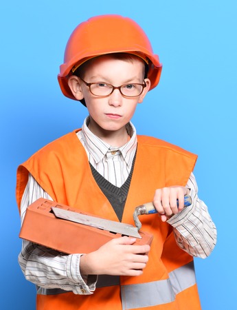 young cute builder boy in orange uniform and hard hat or helmet with glasses holding tool or putty knife and brick on blue studio backgroundの写真素材