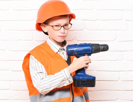 young cute builder boy in orange uniform and hard hat or helmet with glasses holding electric screwdriver on white brick wall backgroundの写真素材