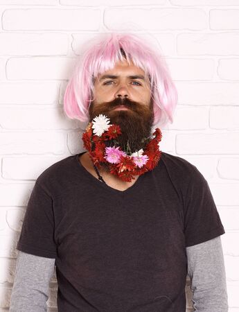 handsome bearded man with stylish mustache in pink wig with colorful autumn flowers in long beard on serious face on white brick wall backgroundの写真素材