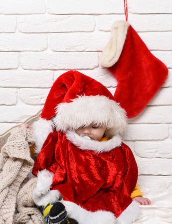 small baby boy with adorable curious face in red sweater with dummy in new year hat and christmas or xmas stocking or boot on white brick wall backgroundの写真素材