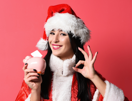 pretty cute  santa girl or smiling brunette woman in new year sweater and christmas or xmas hat holds pink piggy pig bank and showing ok on red studio backgroundの写真素材