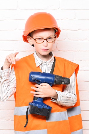 young cute builder boy in orange uniform and hard hat or helmet with glasses holding electric...