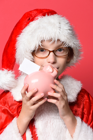 young cute santa claus boy with glasses in xmas sweater and new year christmas hat holding and kissing pink piggy pig bank on red studio backgroundの写真素材