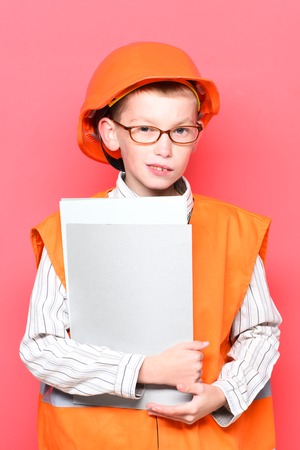 young cute builder boy in orange uniform and hard hat or helmet holding paper on red studio backgroundの写真素材