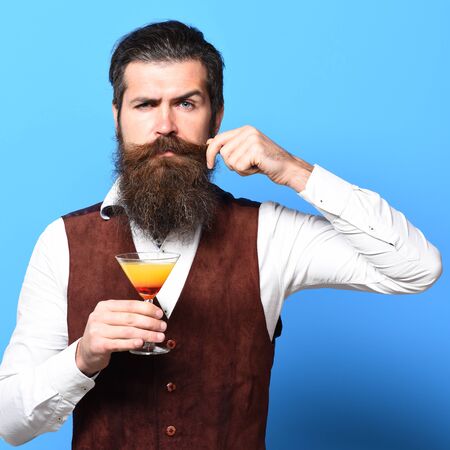 handsome bearded man with long beard and mustache has stylish hair on serious face holding glass of alcoholic cocktail in vintage suede leather waistcoat on blue studio backgroundの写真素材