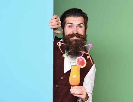 handsome bearded man with in long beard and mustache has stylish hair on smiling face holding glass of alcoholic cocktail in vintage suede leather waistcoat on colorful studio backgroundの写真素材
