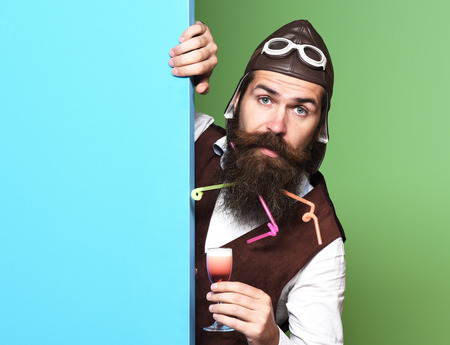 handsome bearded pilot man with long beard and mustache on surprised face holding glass of alcoholic shot in vintage suede leather waistcoat with hat and glasses on blue green studio backgroundの写真素材