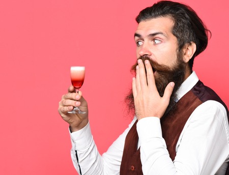 handsome bearded man with long beard and mustache has stylish hair on surprised face holding glass of alcoholic shot in vintage suede leather waistcoat on red studio backgroundの写真素材