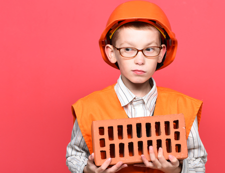 young cute builder boy in orange uniform and hard hat or helmet with glasses holding brick on red studio background, copy spaceの写真素材