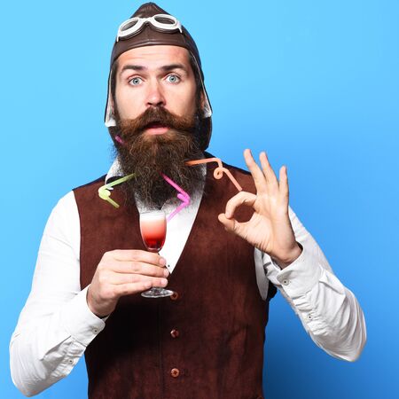 handsome bearded pilot or aviator man with long beard and mustache on funny face holding glass of alcoholic shot in vintage suede leather waistcoat with hat and glasses on blue studio backgroundの写真素材