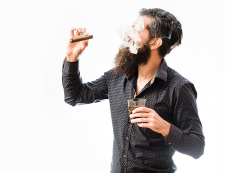 handsome bearded tough rich man with stylish hair mustache and long beard on serious face in blue fashion shirt smoking cigar and holding glass of whiskey isolated on whiteの写真素材
