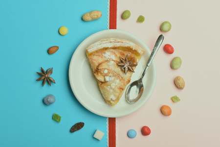 Single slice of apple pie with icing sugar, anise, peanuts, raisins, colorful dragee, marmalade or jelly candies and white plate, spoon, red ribbon on colorful backgroundの写真素材