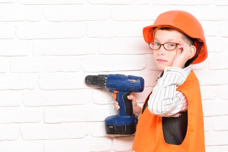 young cute builder boy in orange uniform and hard hat or helmet with glasses holding electric screwdriver on white brick wall background, copy spaceの写真素材