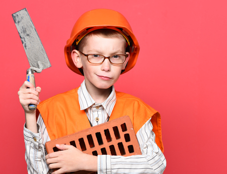 young cute builder boy in orange uniform and hard hat or helmet with glasses holding tool or putty knife and brick on red studio background, copy spaceの写真素材