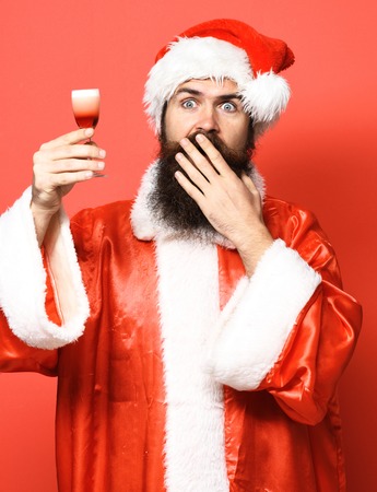 handsome bearded santa claus man with long beard on surprised face holding glass of alcoholic shot in christmas or xmas sweater and new year hat on red studio backgroundの写真素材