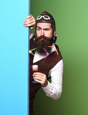 handsome bearded pilot or aviator man with long beard and mustache on serious face holding glass of alcoholic shot in vintage suede leather waistcoat with hat and glasses on colorful studio backgroundの写真素材