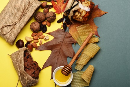 Autumn composition of peanuts, almonds, hazelnuts, brasil nut, walnuts with oatmeal cookies, honey, honeycomb, raisins, knitted bag, maple leaves on colorful backgroundの写真素材