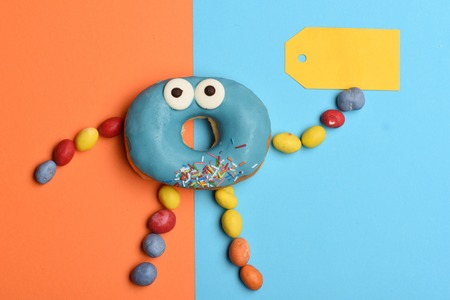 Blue funny surprised glazed donut with icing or sprinkles, colorful dragee with raisins or peanuts inside and yellow tag on colorful backgroundの写真素材