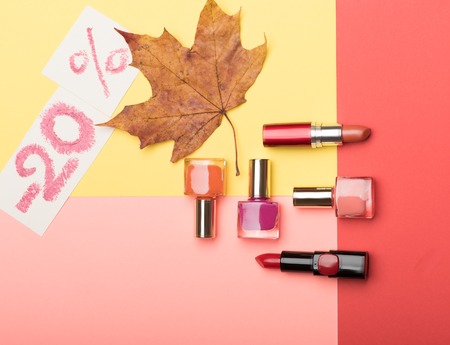 Bottles with nail polish, red lipstick on autumn sale - 20% and maple leaf on colorful backgroundの写真素材