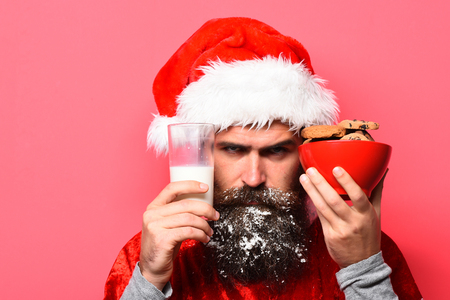 handsome bearded man with stylish mustache and long snowy beard on serious face holding glass of milk with chocolate chip cookies in red santa suit on studio backgroundの写真素材