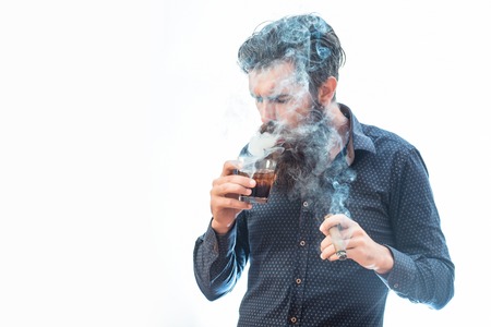 handsome bearded tough rich man with stylish hair mustache and long beard on serious face in blue fashion shirt smoking cigar and holding glass of whiskey isolated on white, copy spaceの写真素材