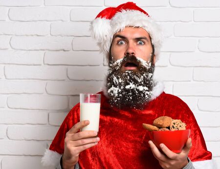 handsome bearded man with stylish mustache and long snowy beard on surprised face holding glass of milk with chocolate chip cookies in red santa suit on white brick wall backgroundの写真素材