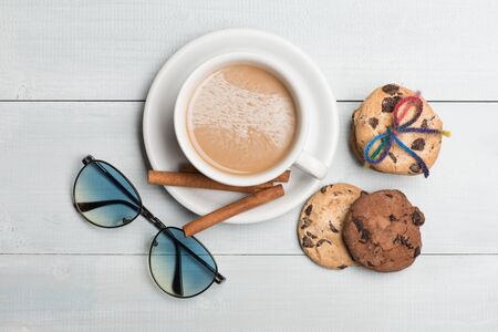White cup of coffee with milk on plate, cinnamon, chocolate chip cookies tied with rainbow string and stylish glasses on vintage wooden background, top viewの写真素材
