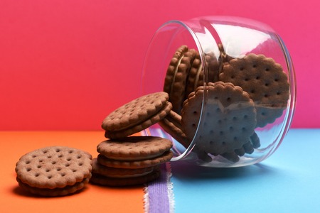Chocolate chip cookies poured from glass jar with purple ribbon on colorful pink orange blue backgroundの写真素材