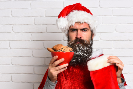 handsome bearded man with stylish mustache and long snowy beard on serious face holding chocolate chip cookies and christmas sock in red santa suit on white brick wall background, copy spaceの写真素材