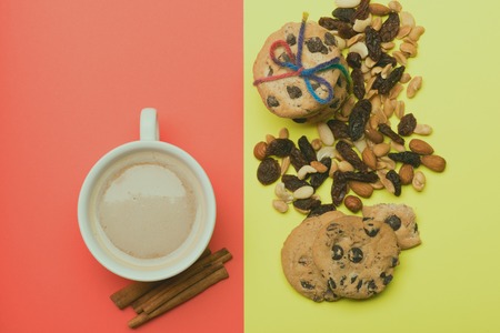 White cup of coffee with milk or cappuccino with chocolate chip cookies tied with rainbow string, different types of nuts and cinnamon on colorful red yellow background, top viewの写真素材
