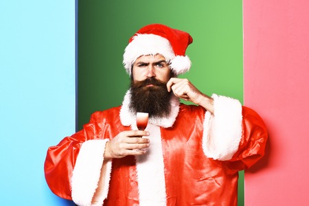handsome bearded santa claus man with long beard on serious face holding glass of alcoholic shot in red christmas or xmas coat and new year hat on colorful studio backgroundの写真素材