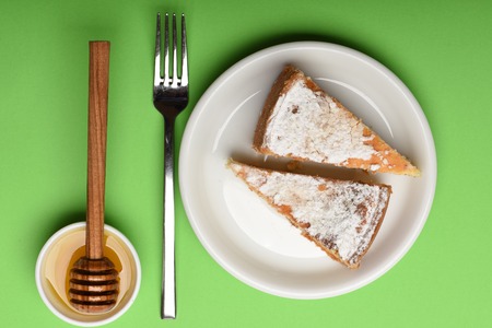tasty slice of apple pies on white plate near silver fork, honey and stick on green background, top...