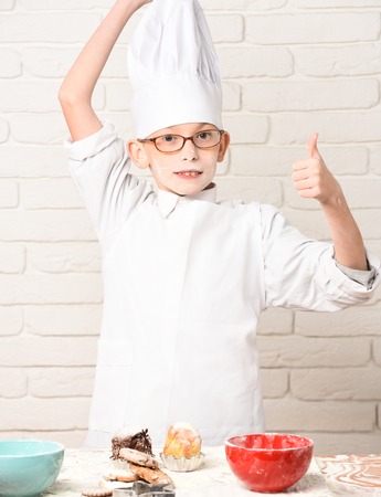 young boy cute cook chef in white uniform and hat on stained face flour with glasses standing near table with rolling pin and colorful bowls with chocolate cookies on brick wall backgroundの写真素材