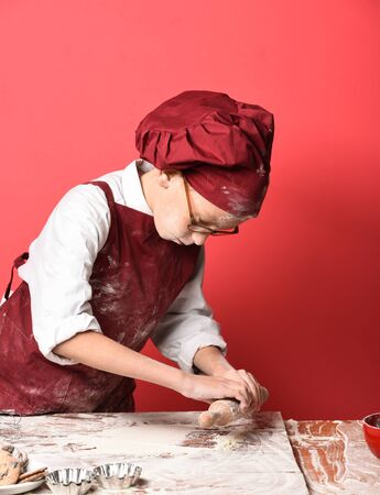 young small boy cute cook chef in uniform and hat on stained face flour with glasses standing near table holding rolling pin and cooking on red studio backgroundの写真素材