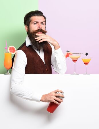 handsome bearded barman with long beard and mustache has stylish hair on serious face holding shaker and made alcoholic cocktail in vintage suede leather waistcoat on purple green studio backgroundの写真素材