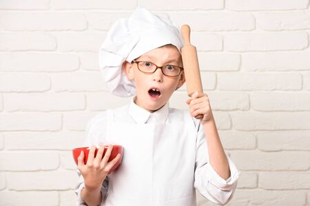 young boy small cute cook chef in white uniform and hat on surprised face with glasses holding red bowl with rolling pin on brick wall backgroundの写真素材