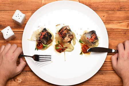 delicious dish of fish with vegetables on white an oval plate and male hands with fork and knife and salt on brown vintage wooden background, top viewの写真素材