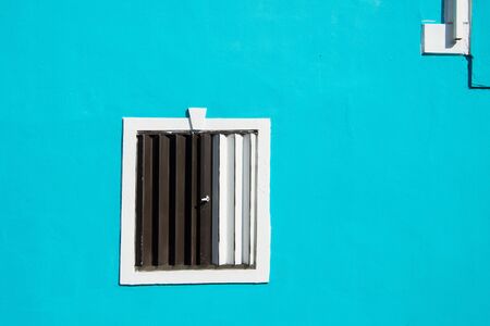 Window with shutters closed and undercolored on plastered facade wall of residential building outdoors on sunny day on turquoise backgroundの写真素材