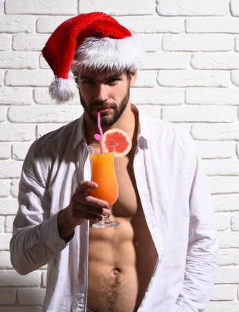 young handsome bearded sexy christmas man with stylish beard in red santa claus hat and white shirt with muscular bare torso holding glass of alcoholic orange beverage on brick wall backgroundの写真素材