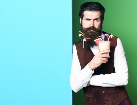 handsome bearded man with long beard and mustache has stylish hair on serious face holding glass of alcoholic beverage in vintage suede leather waistcoat on blue green studio background, copy spaceの写真素材