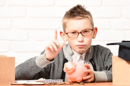 young cute pupil boy in grey sweater and glasses sitting at desk with copybook pink piggy pig bank and graduation cap in classroom with raised finger on white brick wall backgroundの写真素材