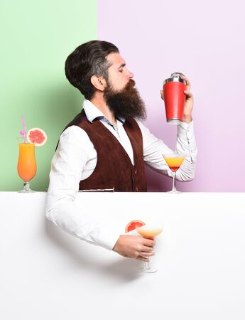 handsome bearded barman with long beard and mustache has stylish hair on serious face holding shaker and made alcoholic cocktail in vintage suede leather waistcoat on purple green studio backgroundの写真素材