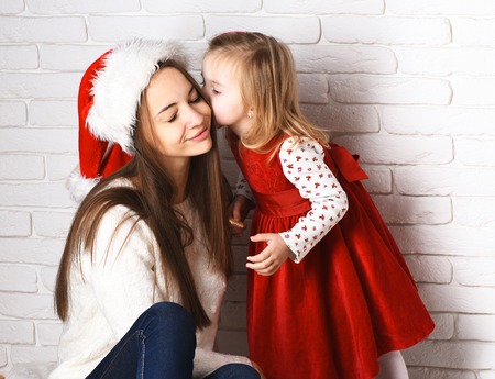 young cute little blonde girl in christmas red dress kissing her smiling mother in xmas hat and long brunette hair on white brick wall backgroundの写真素材